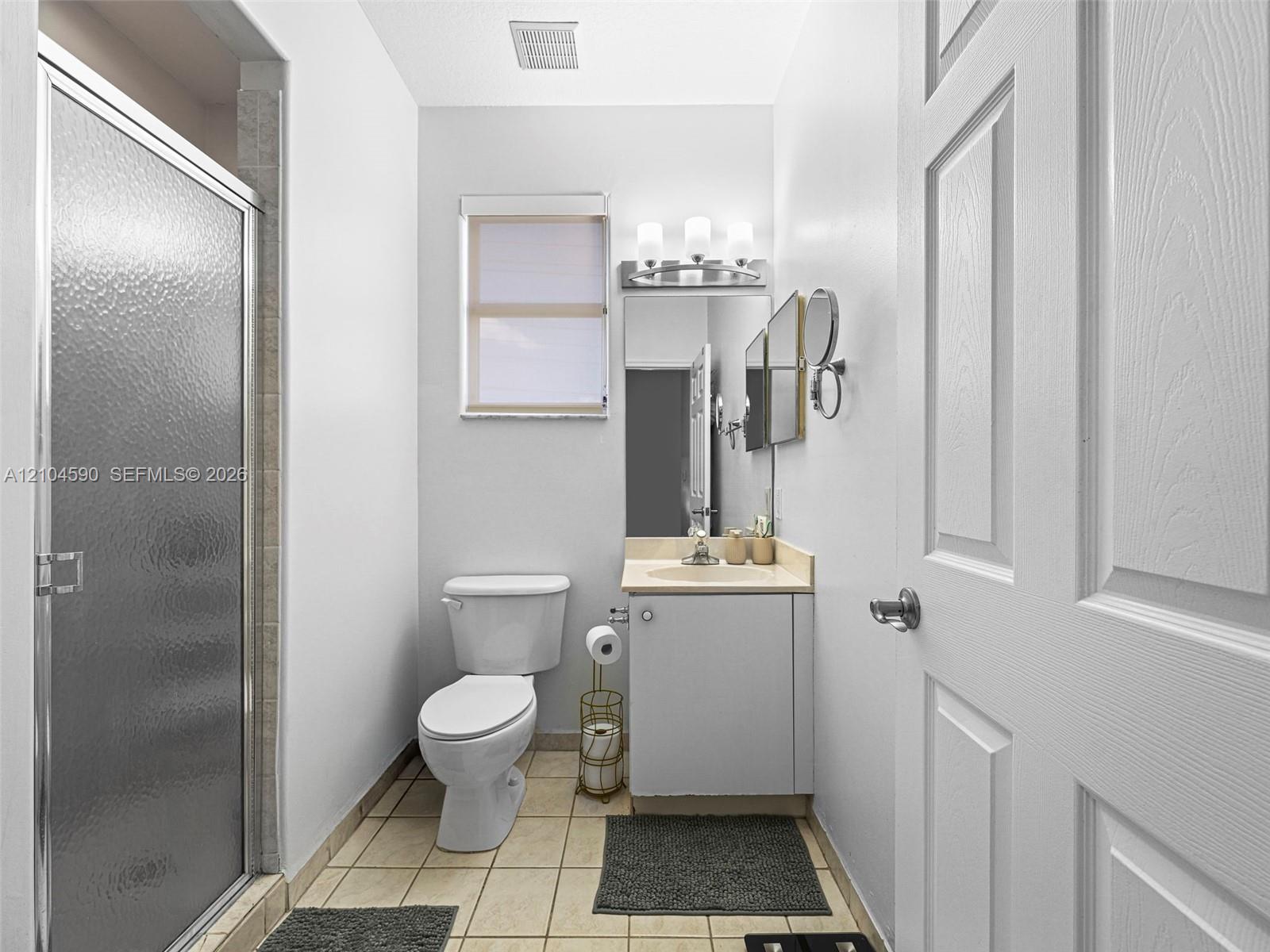 Bathroom featuring a shower stall with a textured glass door, single-basin vanity with a solid surface countertop, rectangular mirror, three-light fixture, and tiled flooring Bathroom featuring a shower stall with a textured glass door, single-basin vanity with a solid surface countertop, rectangular mirror, three-light fixture, and tiled flooring