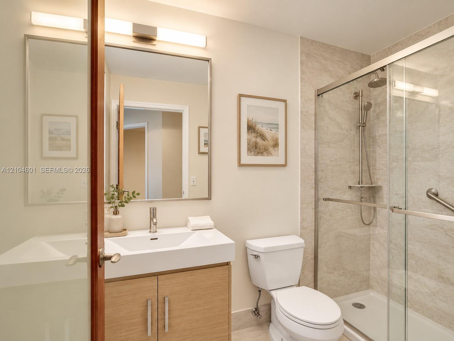Bathroom featuring a wood-finish vanity with a rectangular white sink, frameless glass shower enclosure with neutral stone-style wall tile, wall-mounted brushed-nickel shower fixtures, a rectangular vanity mirror with integrated lighting, and brushed-nickel hardware