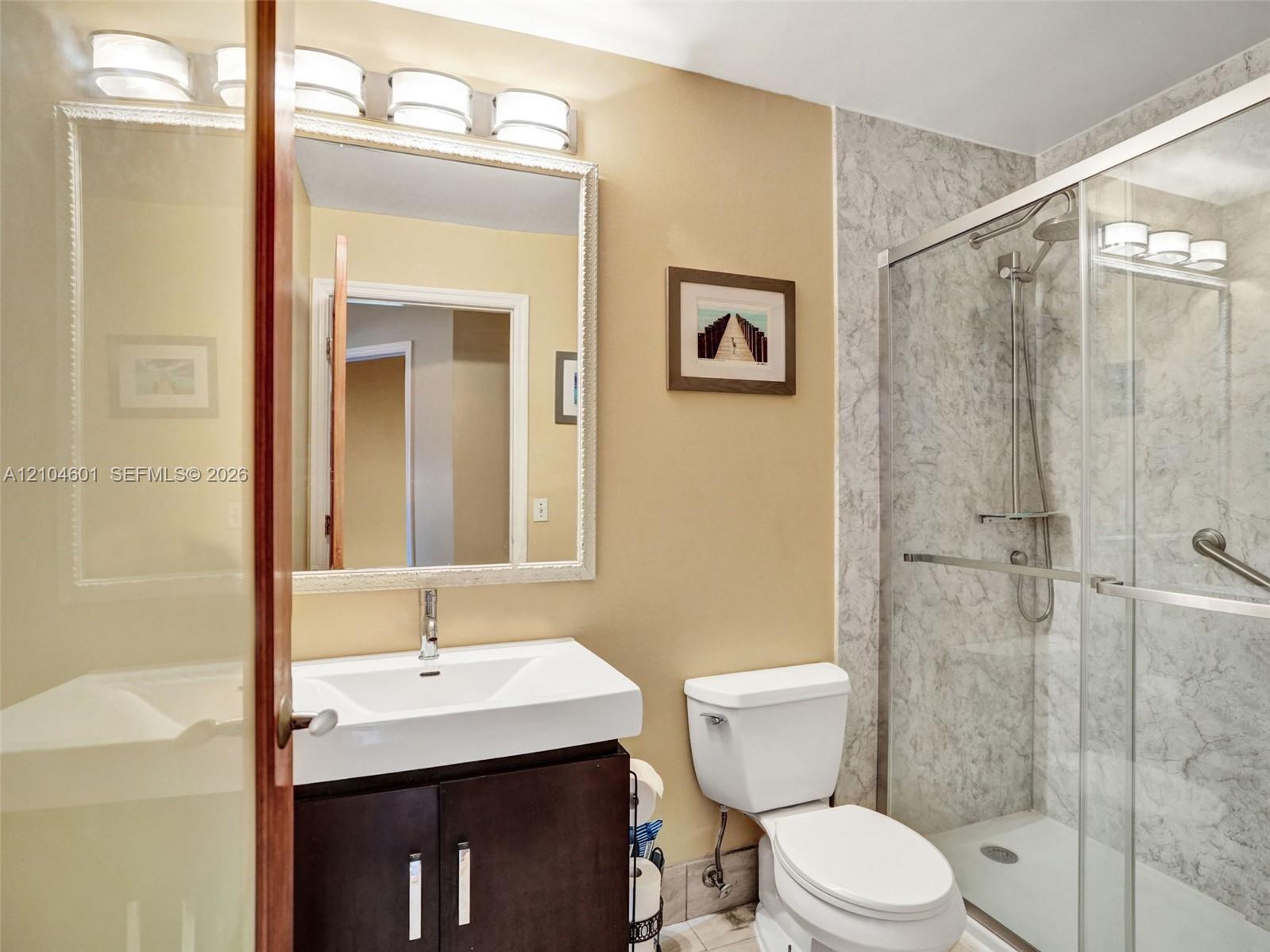 Bathroom featuring a dark-finish vanity with a white rectangular vessel sink, a framed wall mirror, multi-bulb vanity lighting, and a glass-enclosed shower with patterned tile surround