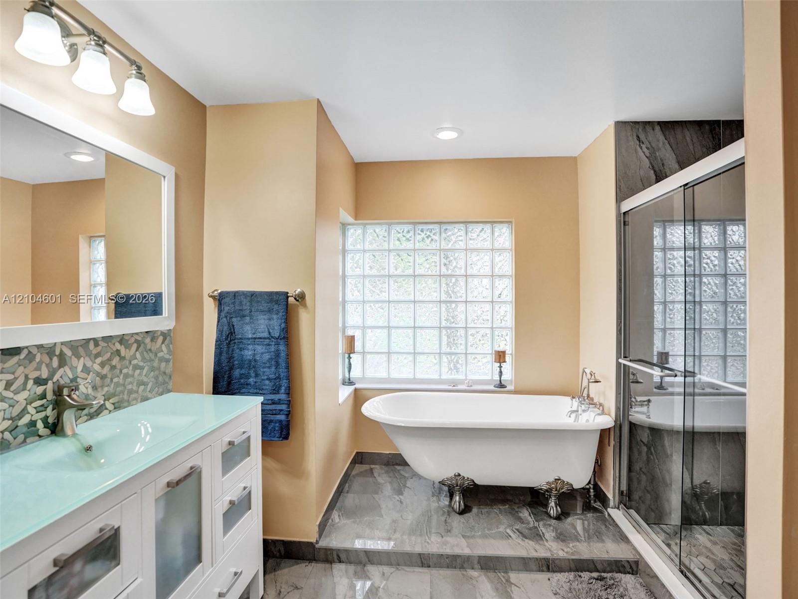 Claw-foot soaking tub, glass-block window, walk-in shower with dark tile surround, integrated glass vanity countertop, and stone-tiled flooring