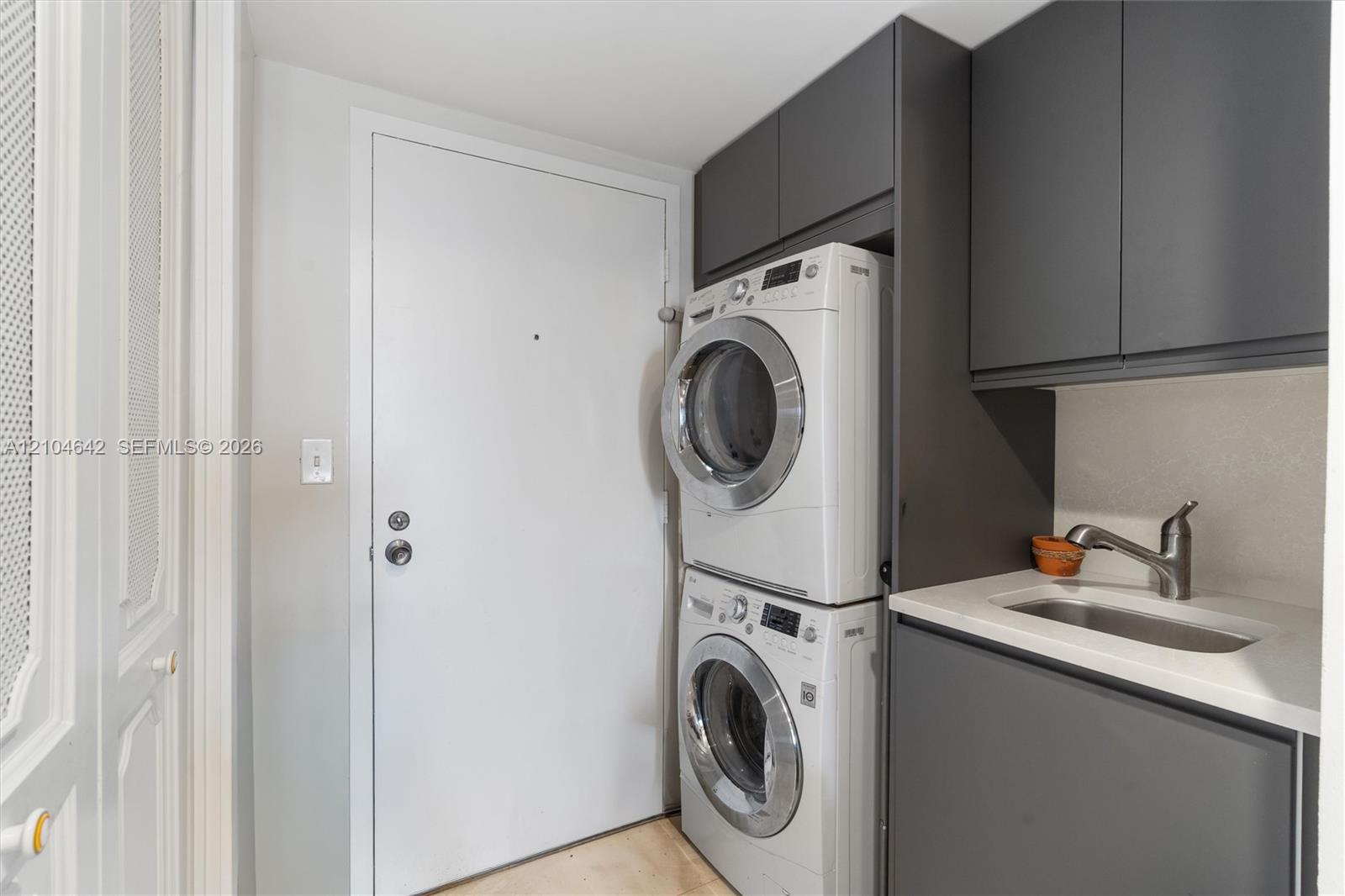 Laundry area featuring charcoal-toned cabinetry, light-toned stone-finish countertop, integrated sink with a gooseneck faucet, and stacked appliance unit