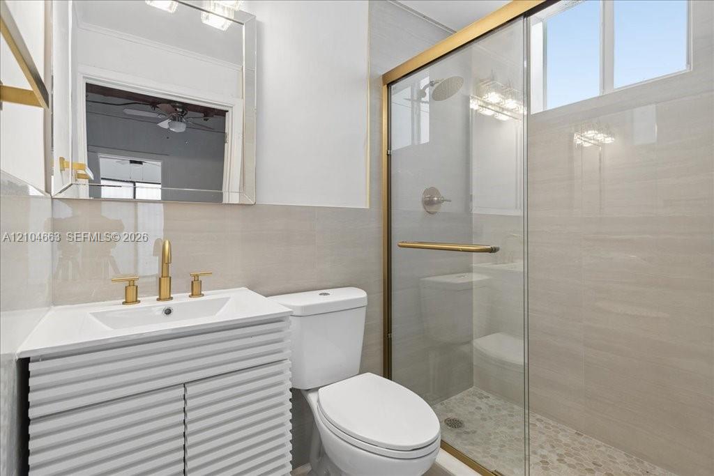 Bathroom featuring a white textured vanity cabinet, gold-tone fixtures, a walk-in shower with a glass enclosure, and light-toned wall tile Bathroom featuring a white textured vanity cabinet, gold-tone fixtures, a walk-in shower with a glass enclosure, and light-toned wall tile