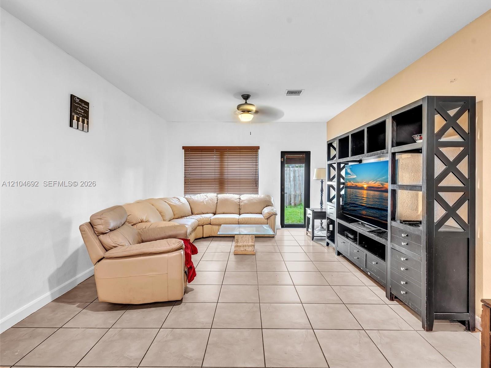 Spacious living area featuring large-format ceramic tile flooring, built-in floor-to-ceiling wooden shelving unit, ceiling fan with integrated lighting, and horizontal window blinds Spacious living area featuring large-format ceramic tile flooring, built-in floor-to-ceiling wooden shelving unit, ceiling fan with integrated lighting, and horizontal window blinds