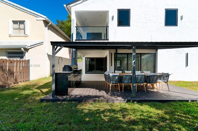 Stucco exterior featuring a covered patio with a dark wood-finish deck, integrated outdoor kitchen, and a glass-railed upper-level balcony