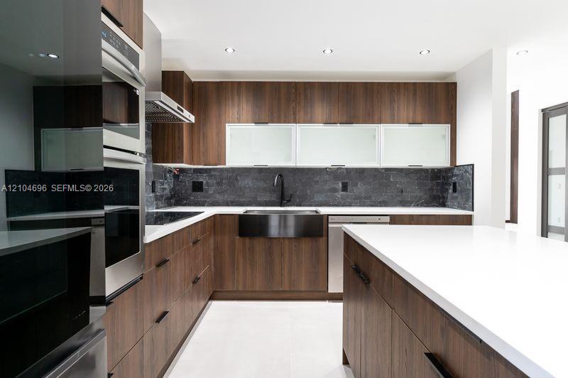 Kitchen featuring wood-finish cabinetry, white countertops, a central island, integrated stainless steel appliances, and a dark stone tile backsplash Kitchen featuring wood-finish cabinetry, white countertops, a central island, integrated stainless steel appliances, and a dark stone tile backsplash