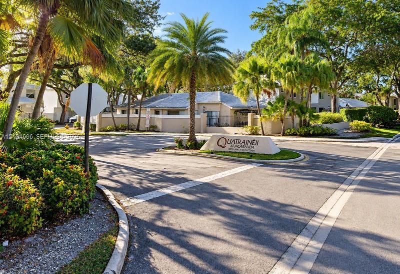 Community entrance featuring paved roadways, mature palm trees, professional landscaping, and a masonry identification monument