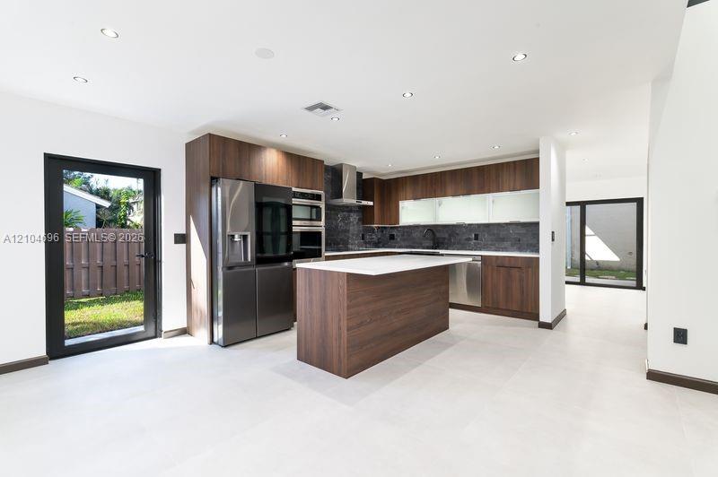 Kitchen featuring flat-panel wood-finish cabinetry, a central island with a solid-surface countertop, integrated stainless steel appliances, dark stone-style backsplash, and recessed lighting Kitchen featuring flat-panel wood-finish cabinetry, a central island with a solid-surface countertop, integrated stainless steel appliances, dark stone-style backsplash, and recessed lighting