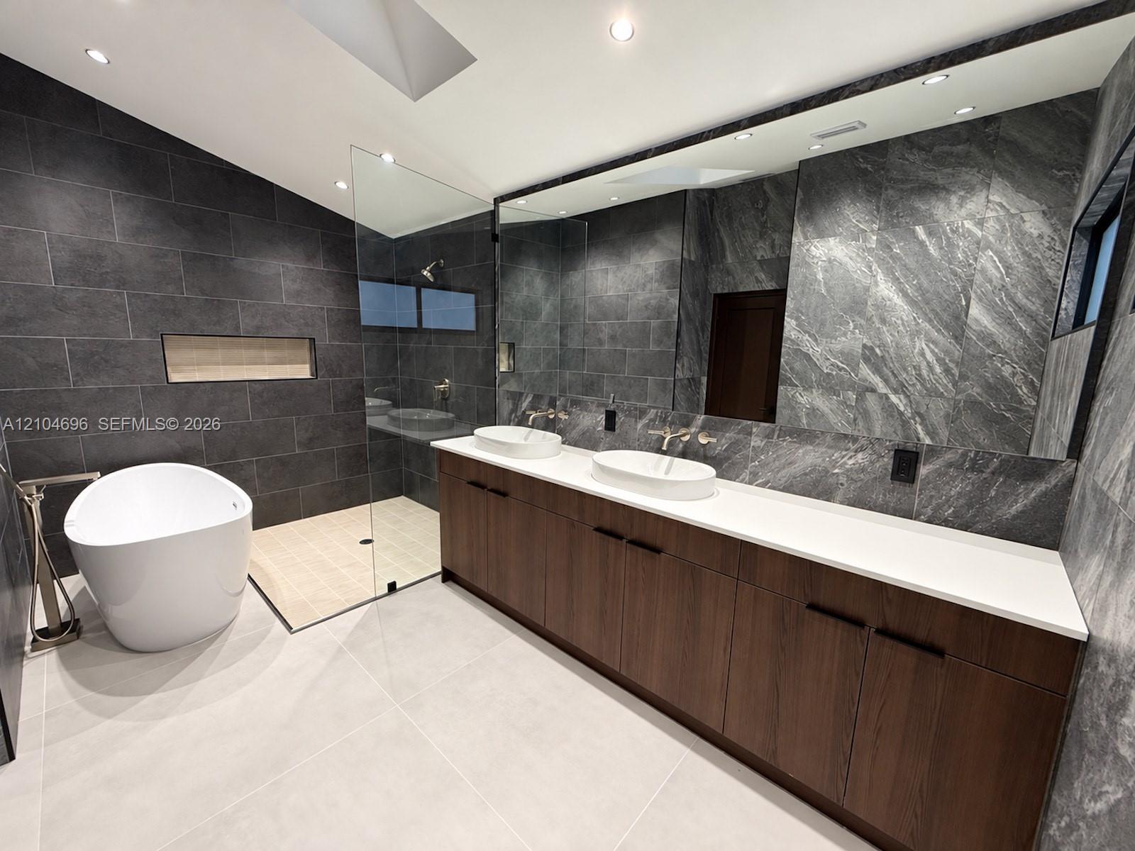 Double vanity with a white solid-surface countertop and dual vessel sinks, freestanding soaking tub, walk-in shower with glass enclosure, wall-mounted matte metal fixtures, and large-format tile wall treatments