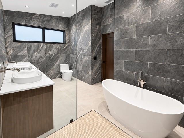 Freestanding white soaking tub with wall-mounted polished chrome faucet, dual vessel sinks atop a wood-finish vanity, and floor-to-ceiling textured stone wall cladding