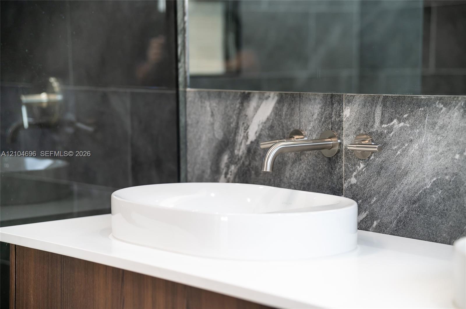 White vessel sink featuring a countertop-mounted vanity with a solid surface top, wall-mounted brushed metal faucet, and large-format tile backsplash