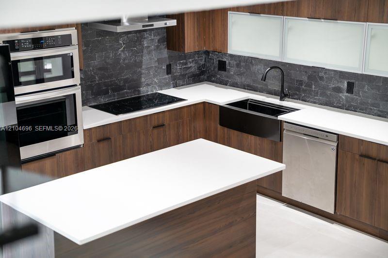 Kitchen featuring wood-finish cabinetry, white countertops, a dark stone backsplash, a black farmhouse-style sink with a matte black faucet, and stainless steel built-in appliances Kitchen featuring wood-finish cabinetry, white countertops, a dark stone backsplash, a black farmhouse-style sink with a matte black faucet, and stainless steel built-in appliances