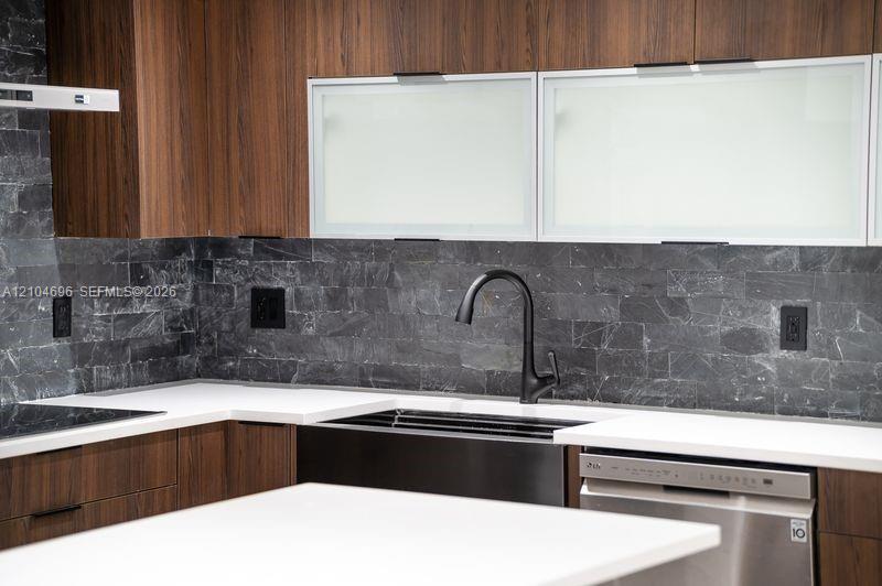Kitchen featuring wood-finish cabinetry, matte black gooseneck faucet, white solid-surface countertops, textured dark stone backsplash, and frosted glass-panel upper cabinet doors Kitchen featuring wood-finish cabinetry, matte black gooseneck faucet, white solid-surface countertops, textured dark stone backsplash, and frosted glass-panel upper cabinet doors