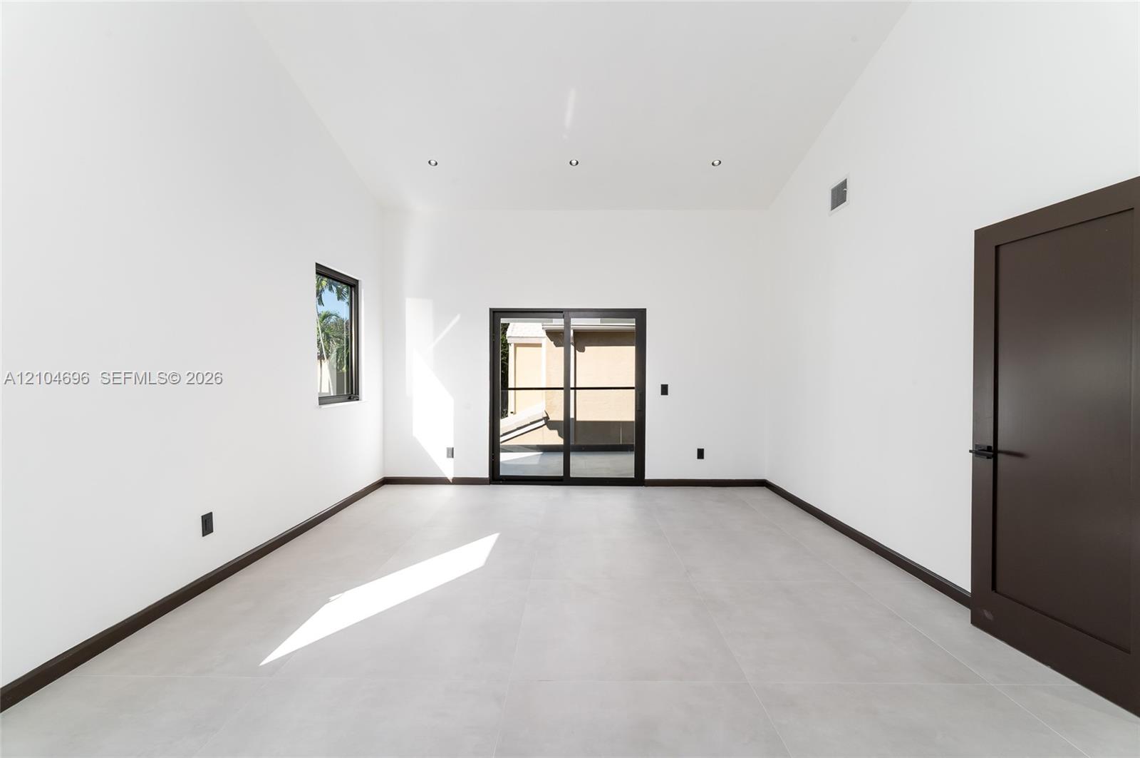 High-ceiling interior featuring large-format tile flooring, recessed lighting, dark-framed sliding glass doors, and a solid-core door with matte black hardware