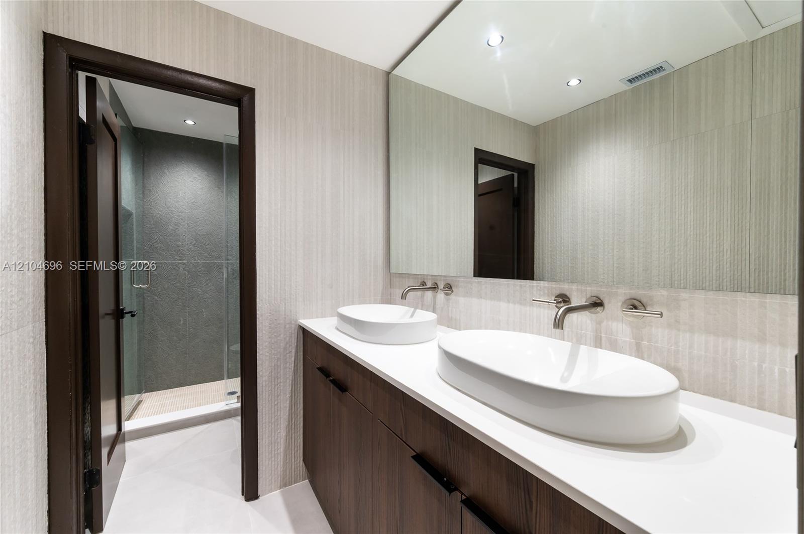 Floating vanity featuring a dual-sink countertop, vessel sinks, wall-mounted faucets, large frameless mirror, and a walk-in shower with glass enclosure