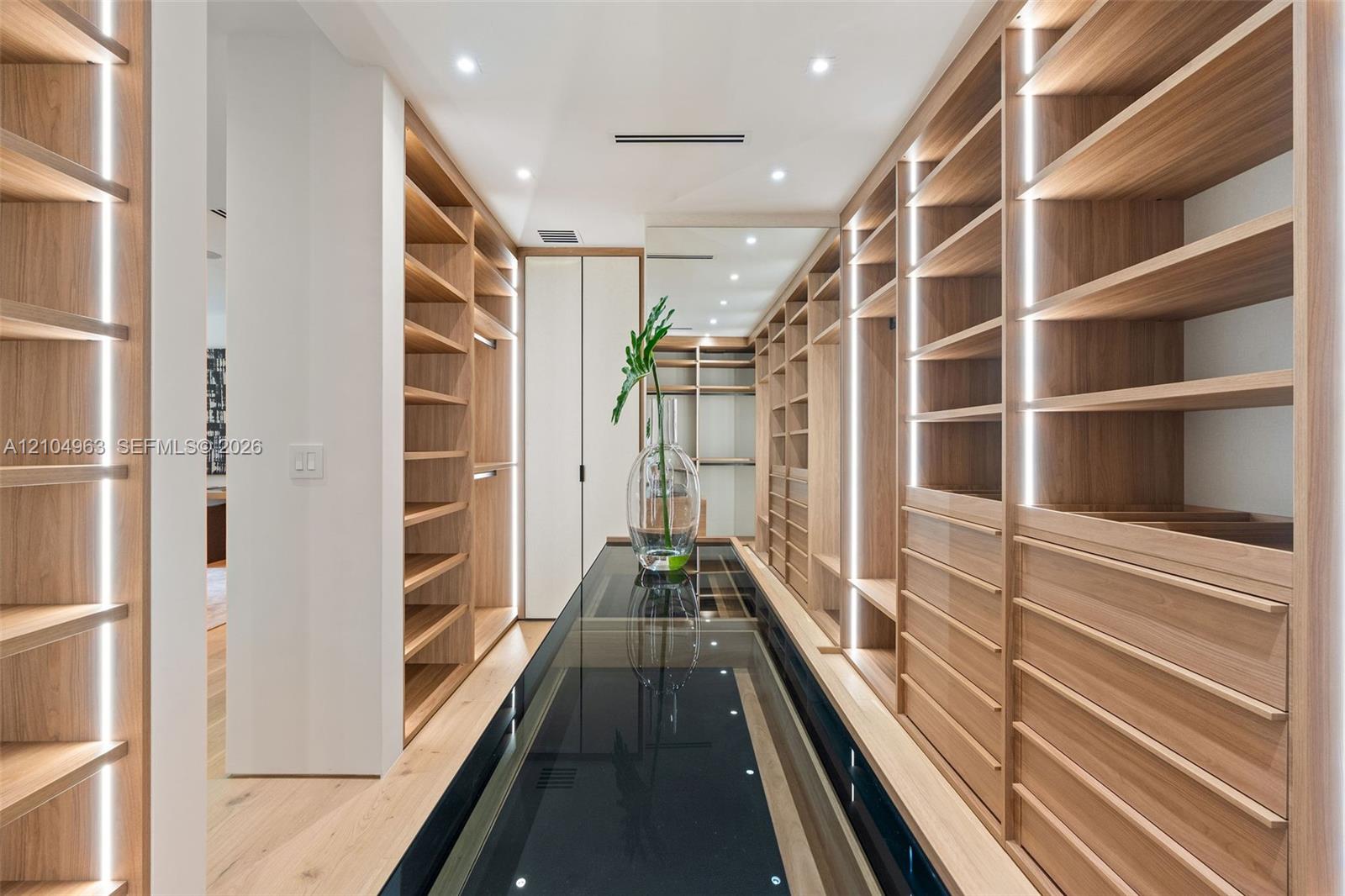 Custom storage cabinetry featuring light wood-finish shelving, integrated vertical LED lighting, a glass-topped center island, and recessed ceiling lighting