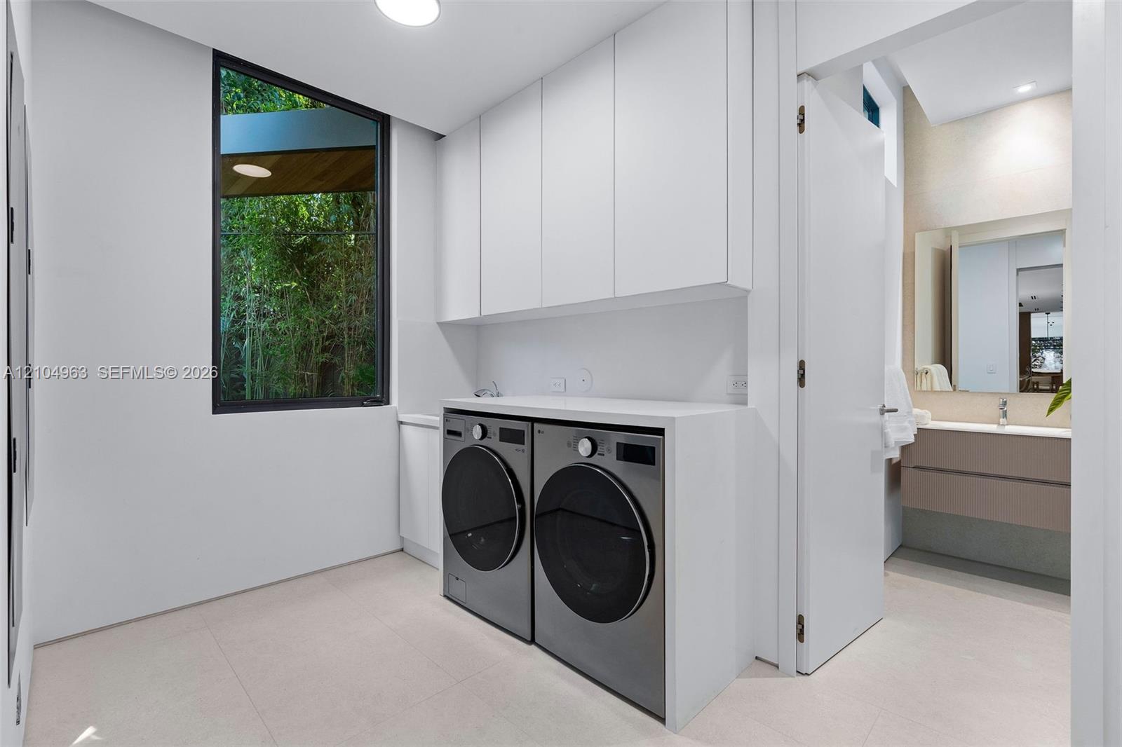 Laundry room featuring floor-to-ceiling cabinetry, custom solid-surface countertops, large-format tile flooring, and a window with natural foliage exposure