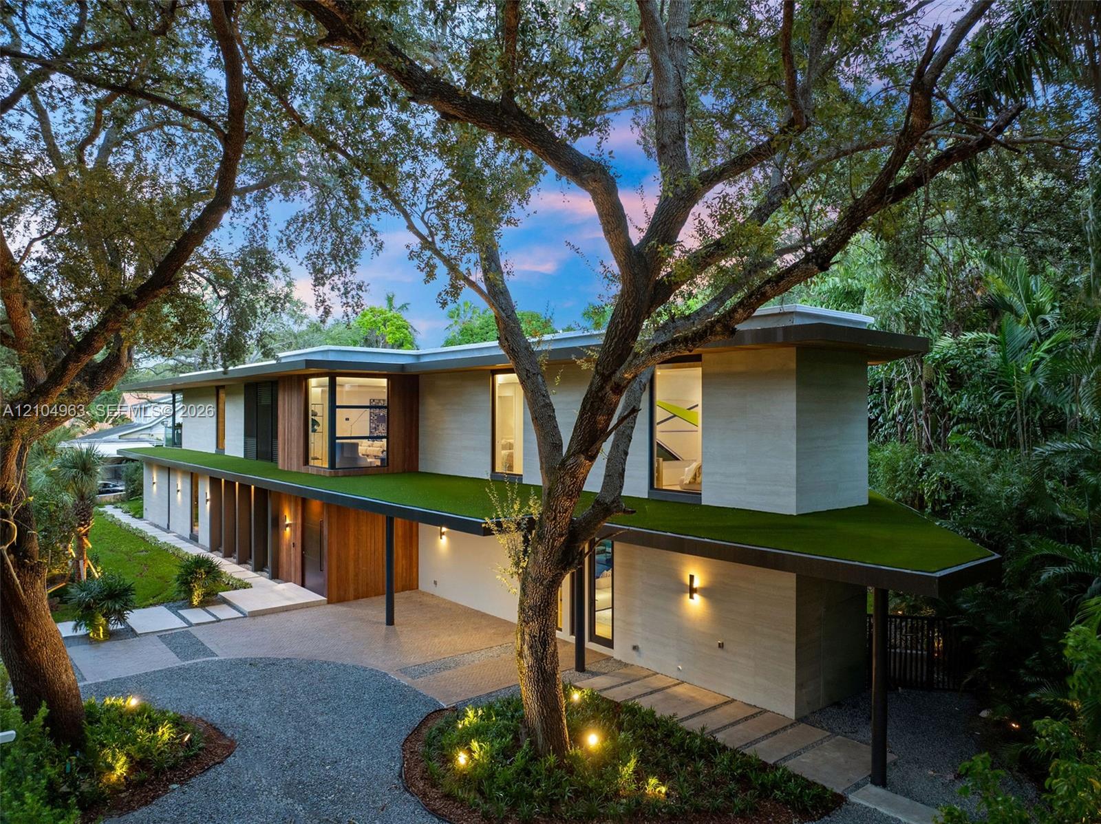Modern architectural exterior featuring a flat roofline, horizontal stone and wood siding, integrated green roof canopy, gravel motor court, and linear concrete pavers