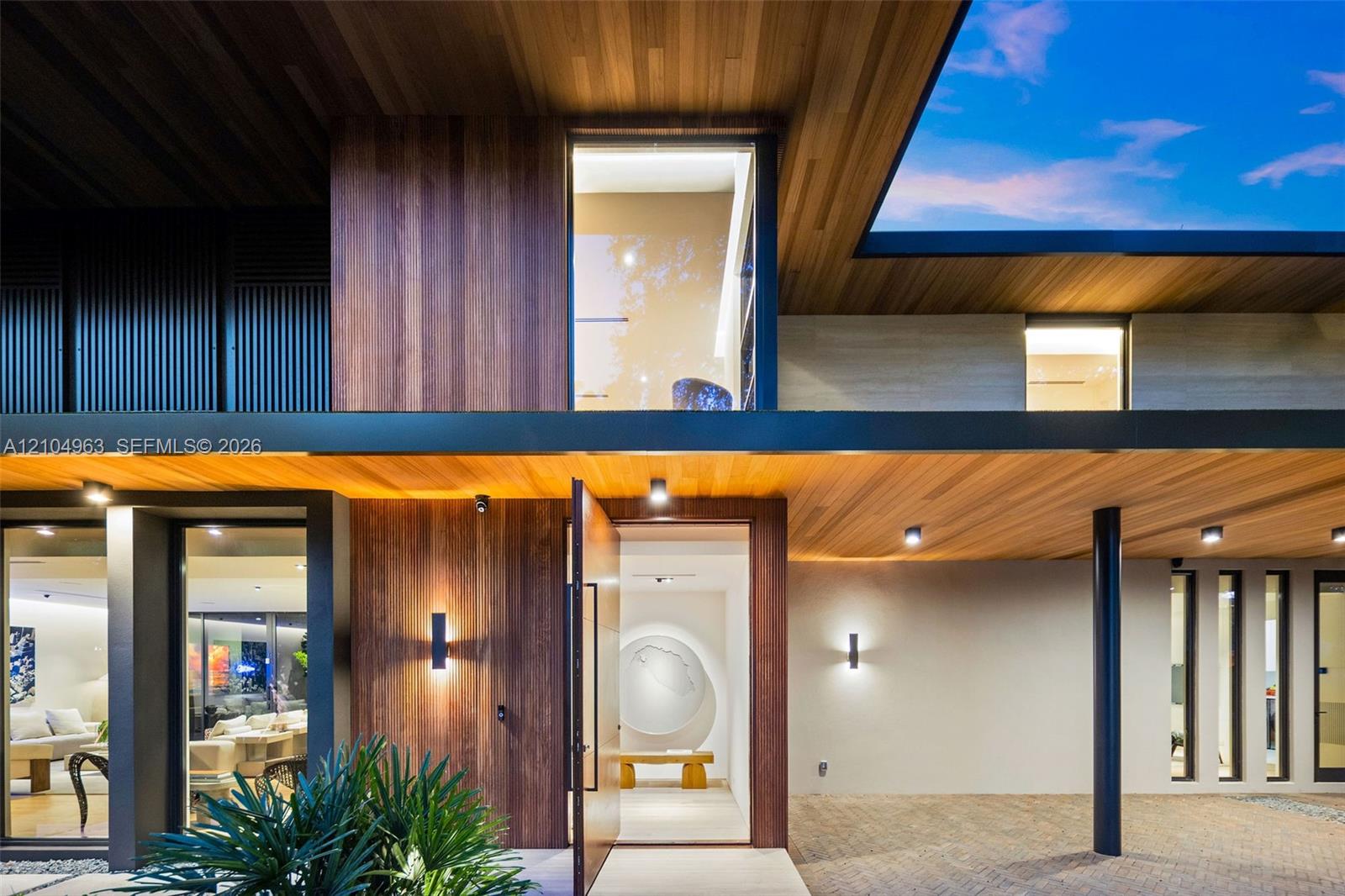 Contemporary facade featuring extensive wood-paneled cladding, floor-to-ceiling glass windows, recessed ceiling lighting, a wide pivot entry door, and covered stone paver driveway Contemporary facade featuring extensive wood-paneled cladding, floor-to-ceiling glass windows, recessed ceiling lighting, a wide pivot entry door, and covered stone paver driveway