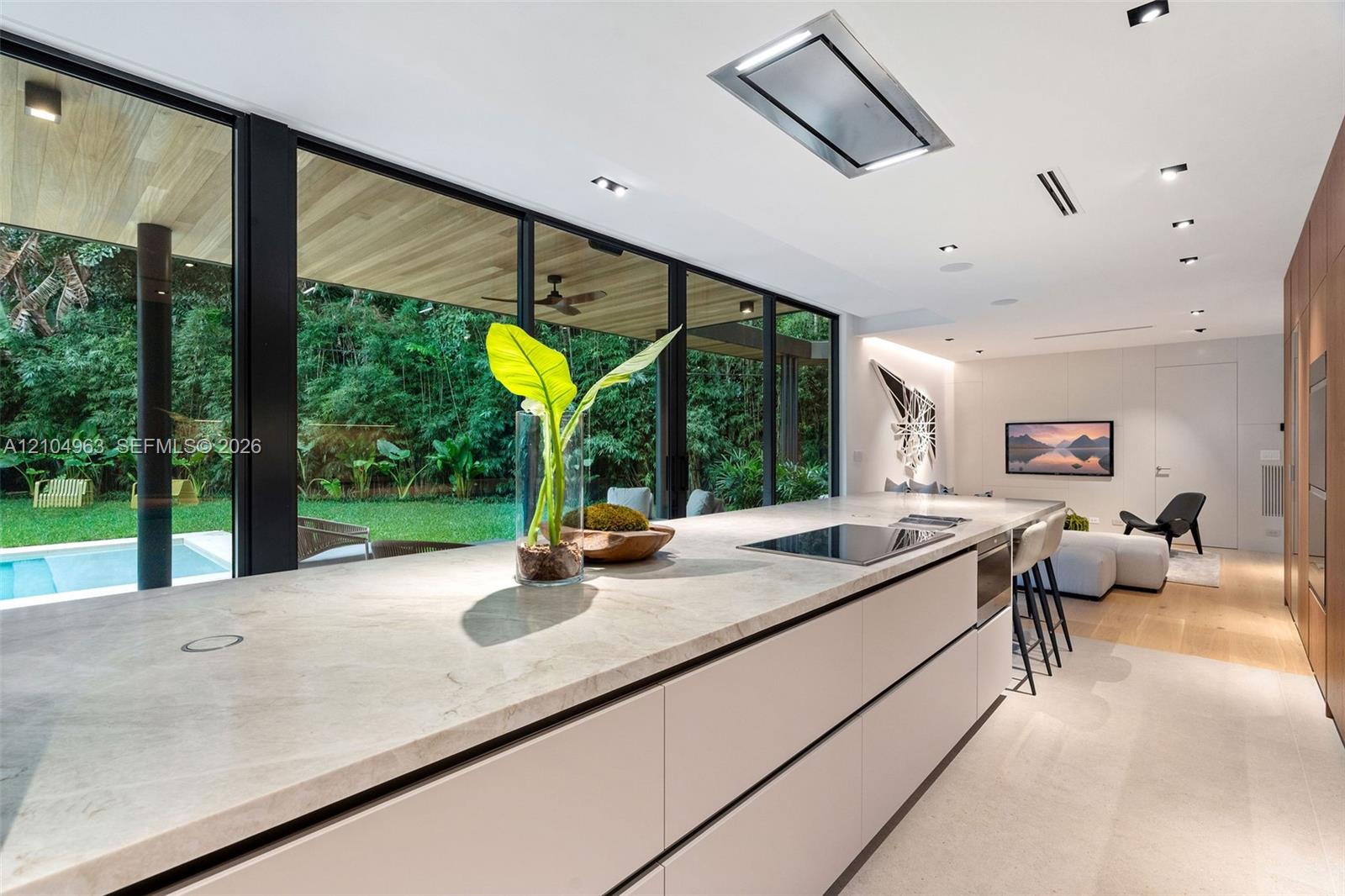 Modern kitchen featuring a stone-topped island, floor-to-ceiling glass panel walls, integrated ceiling lighting, and wood-finish cabinetry
