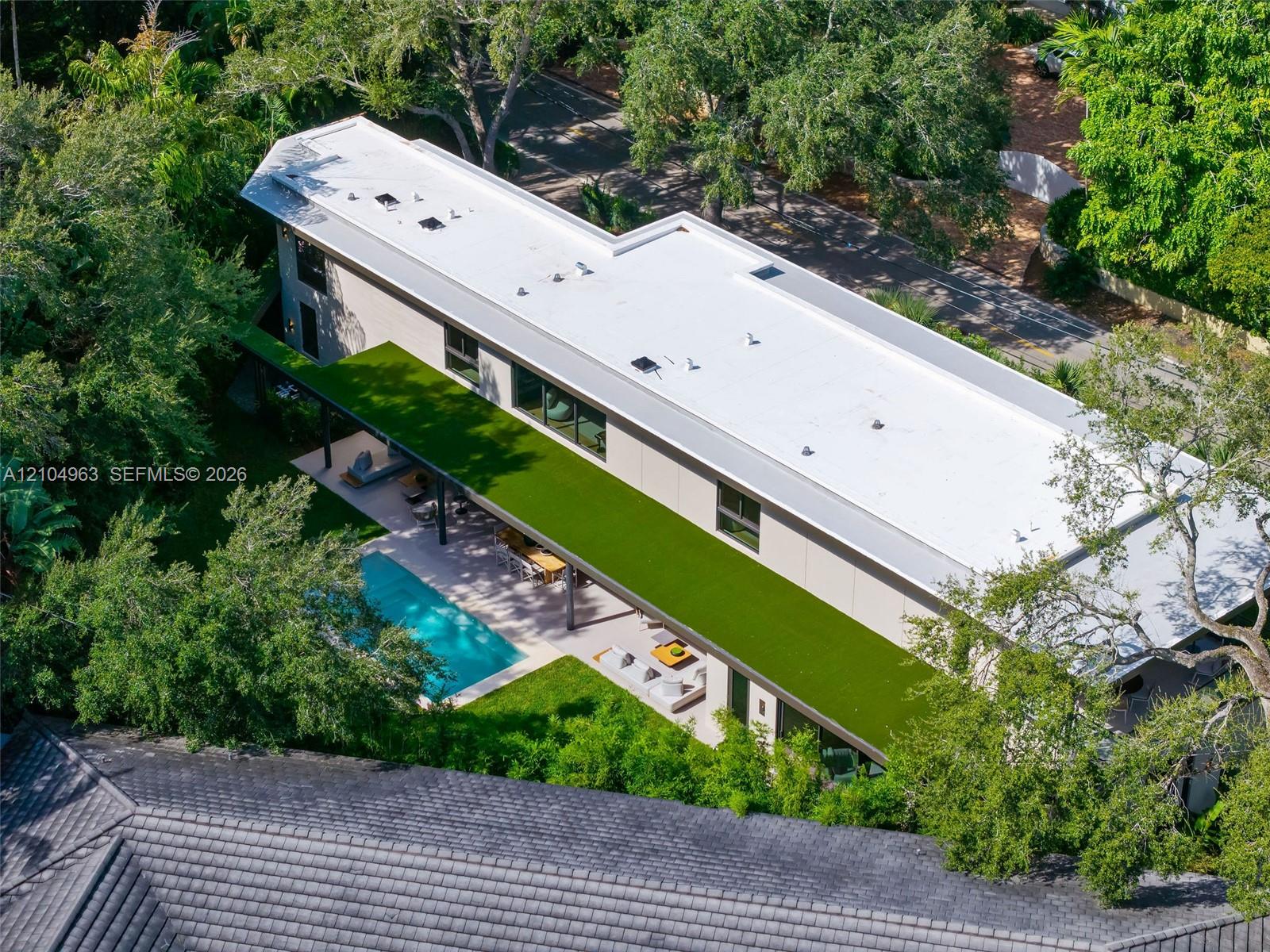 Modern residence featuring a flat roof with integrated skylights, a sprawling covered terrace with an artificial turf canopy, and a private swimming pool set within a manicured backyard