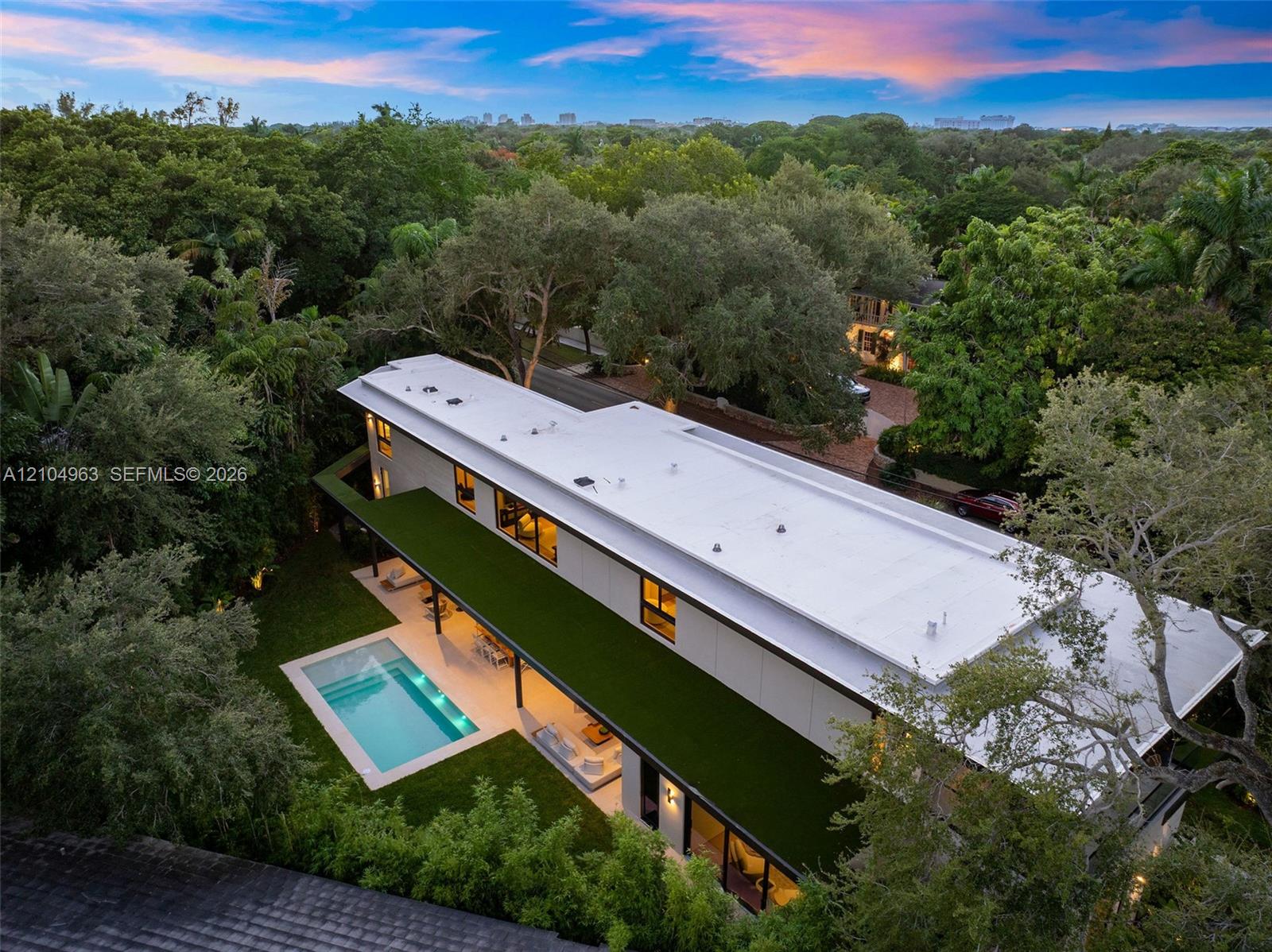 Modern two-story structure featuring a flat roofline, minimalist facade, in-ground swimming pool, expansive hardscaped patio, and mature perimeter landscaping