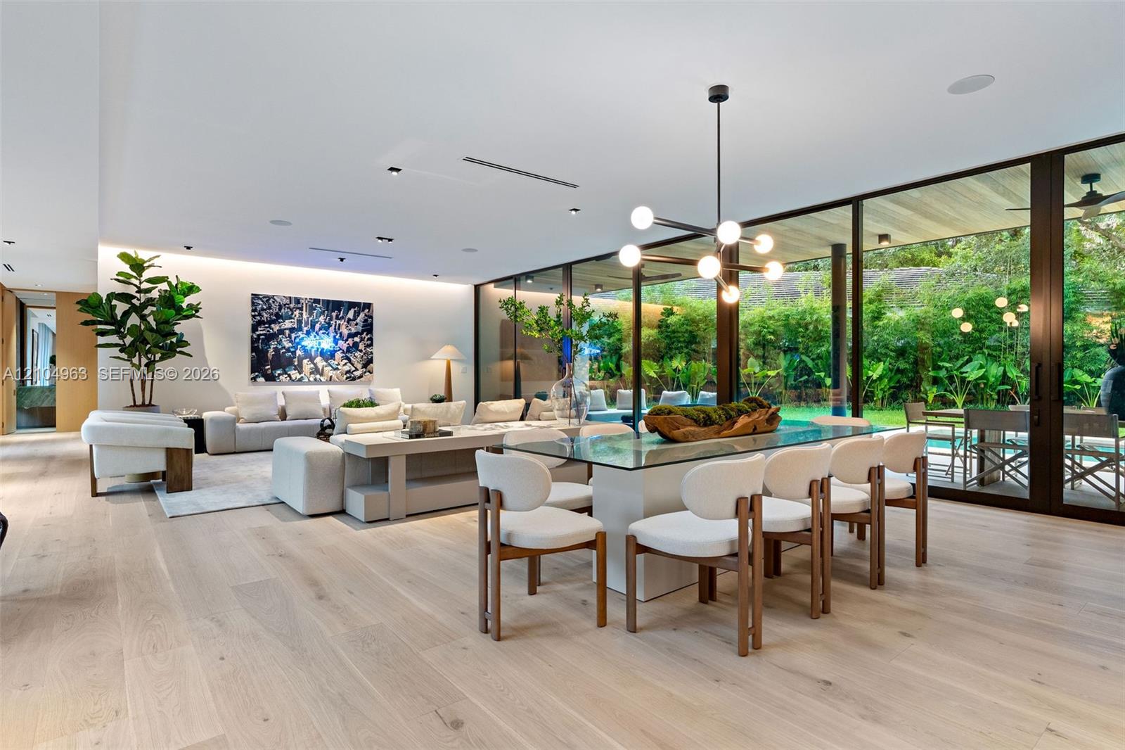 Open-concept interior featuring light wood-finish flooring, floor-to-ceiling glass panel walls, recessed architectural lighting, and a modern multi-arm chandelier Open-concept interior featuring light wood-finish flooring, floor-to-ceiling glass panel walls, recessed architectural lighting, and a modern multi-arm chandelier
