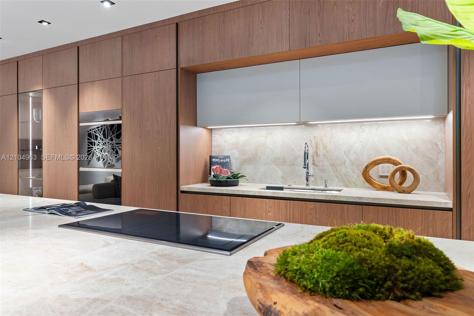 Modern kitchen featuring floor-to-ceiling wood-finish cabinetry, a large stone-surface island with an integrated induction cooktop, light-toned stone backsplash, and recessed ceiling lighting
