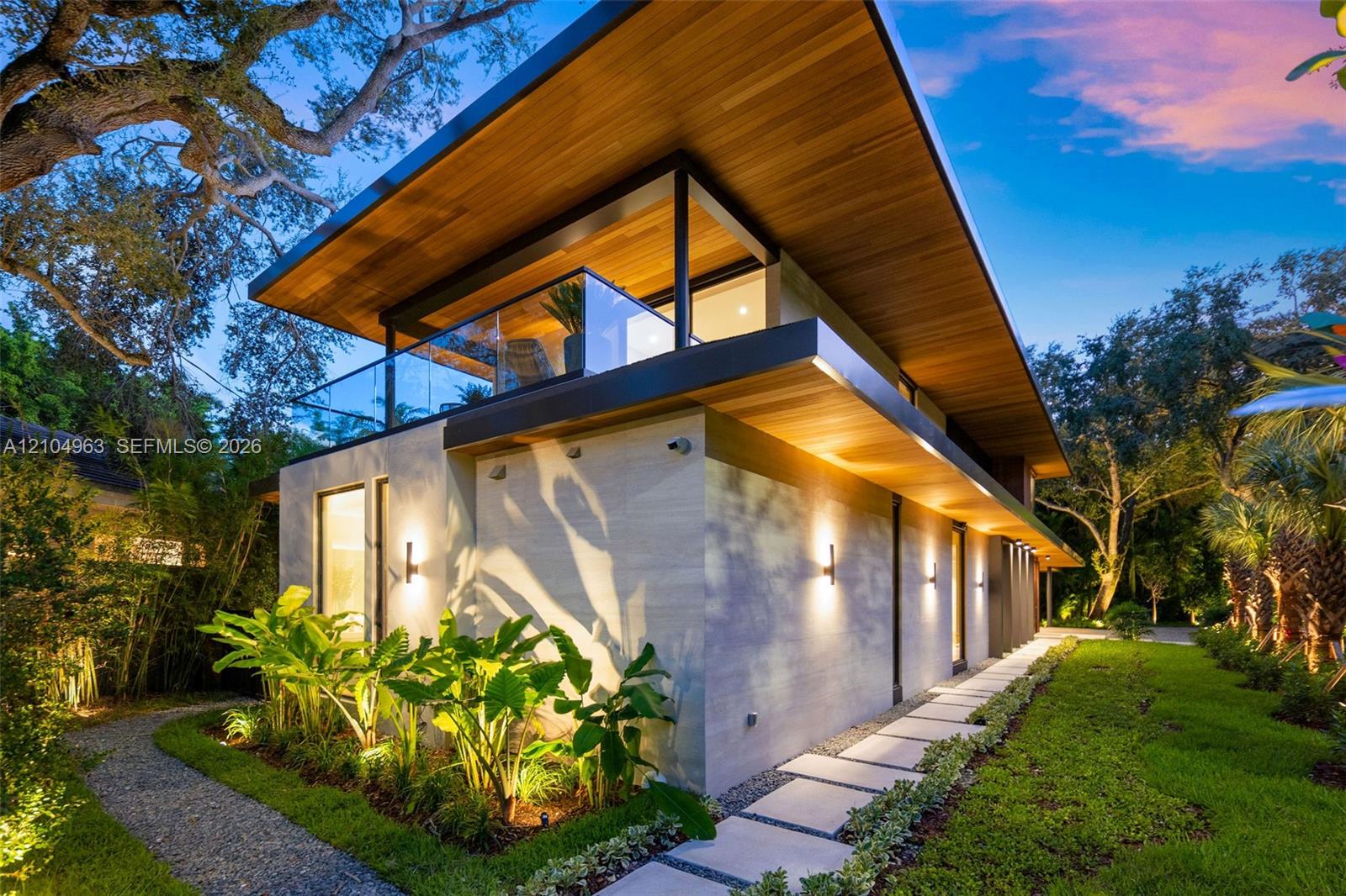 Modern architectural exterior featuring a wood-paneled cantilevered roof, stone-clad facade, glass-paneled balcony railing, and paver stone walkway Modern architectural exterior featuring a wood-paneled cantilevered roof, stone-clad facade, glass-paneled balcony railing, and paver stone walkway