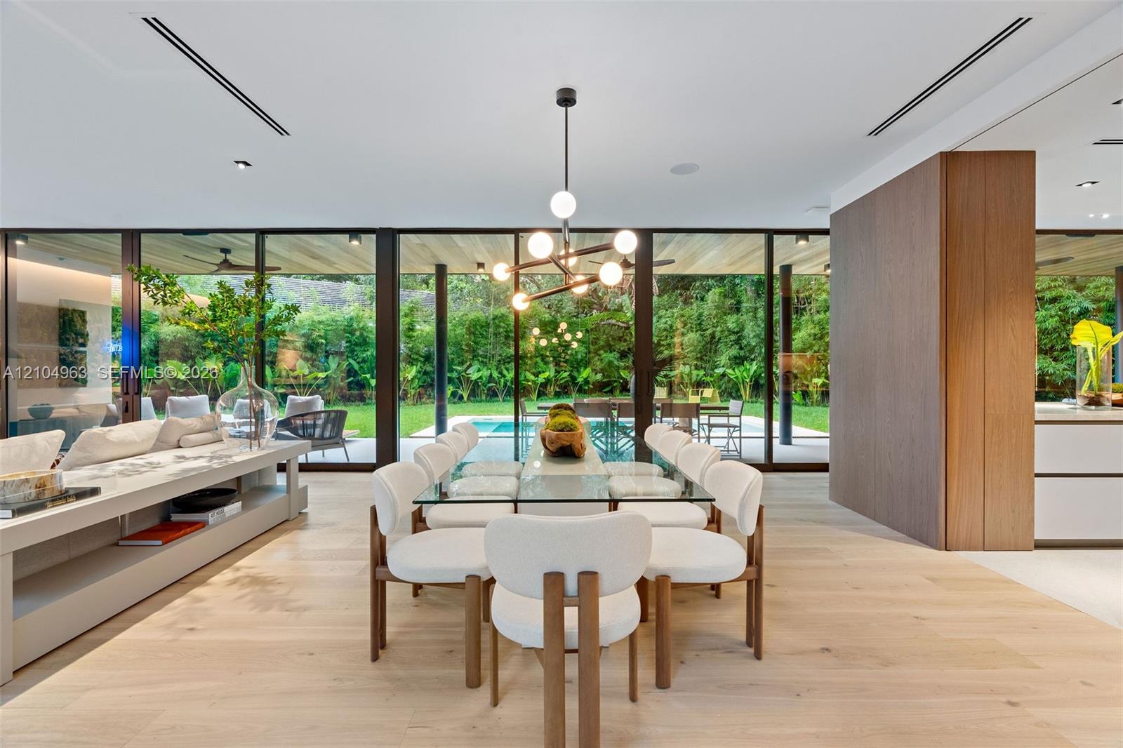 Floor-to-ceiling glass walls, wood-finish flooring, linear recessed lighting, integrated wall cabinetry, and a contemporary multi-arm chandelier Floor-to-ceiling glass walls, wood-finish flooring, linear recessed lighting, integrated wall cabinetry, and a contemporary multi-arm chandelier
