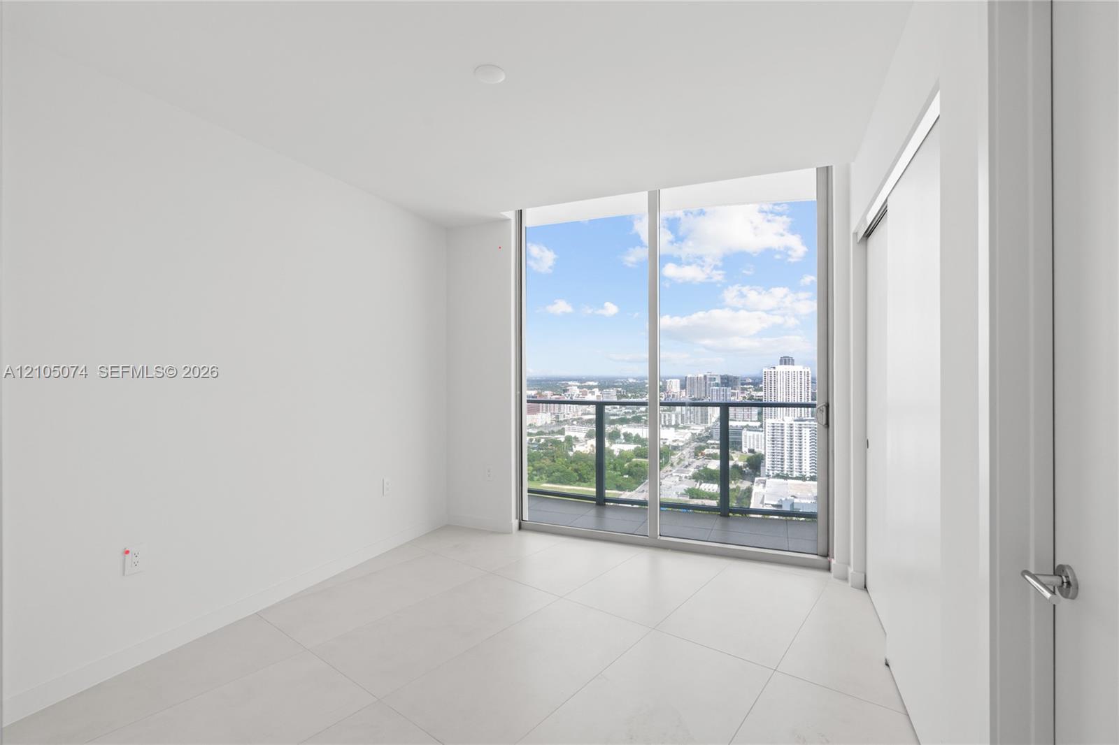 Floor-to-ceiling glass sliding doors, private balcony with metal railing, light-colored tile flooring, and urban horizon scenery