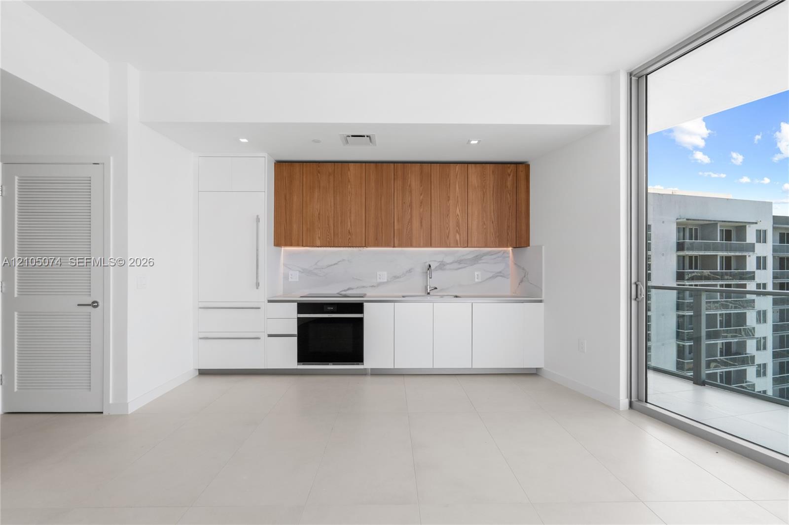 Kitchen featuring floor-to-ceiling cabinetry with wood-finish upper units and white lower cabinetry, stone-patterned backsplash, built-in wall oven, and expansive floor-to-ceiling glass paneling