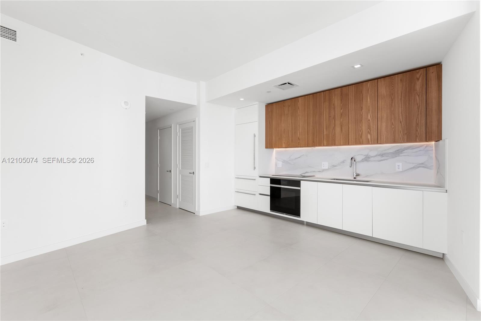 Kitchen featuring flat-panel white and wood-finish cabinetry, integrated appliances, stone-patterned backsplash, and light-toned tile flooring