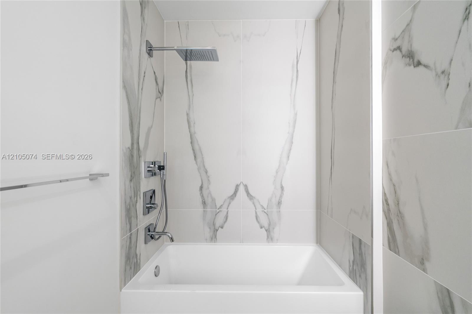 Bathtub featuring large-format veined tile surround, overhead rain shower head, handheld shower wand, and polished chrome fixtures