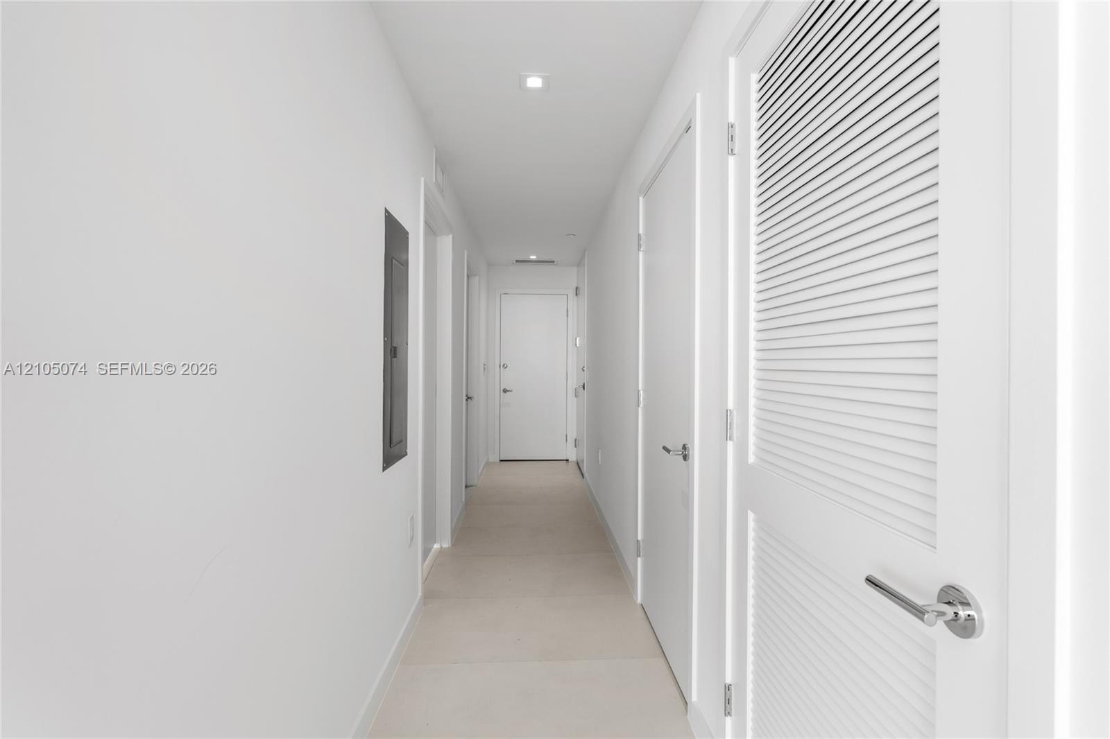 Hallway featuring neutral-toned tile flooring, recessed ceiling lighting, and solid core doors with brushed metal hardware, including a louvered closet door