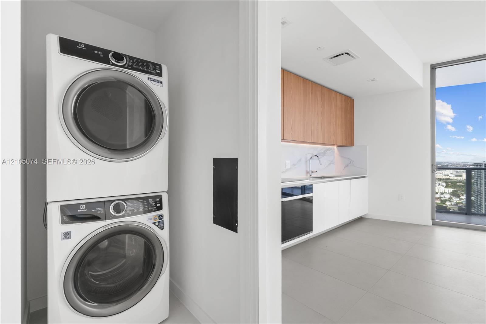 Stacked front-load laundry unit, kitchen featuring white flat-panel cabinetry, wood-finish upper cabinets, integrated stainless steel appliances, and large-format tile flooring