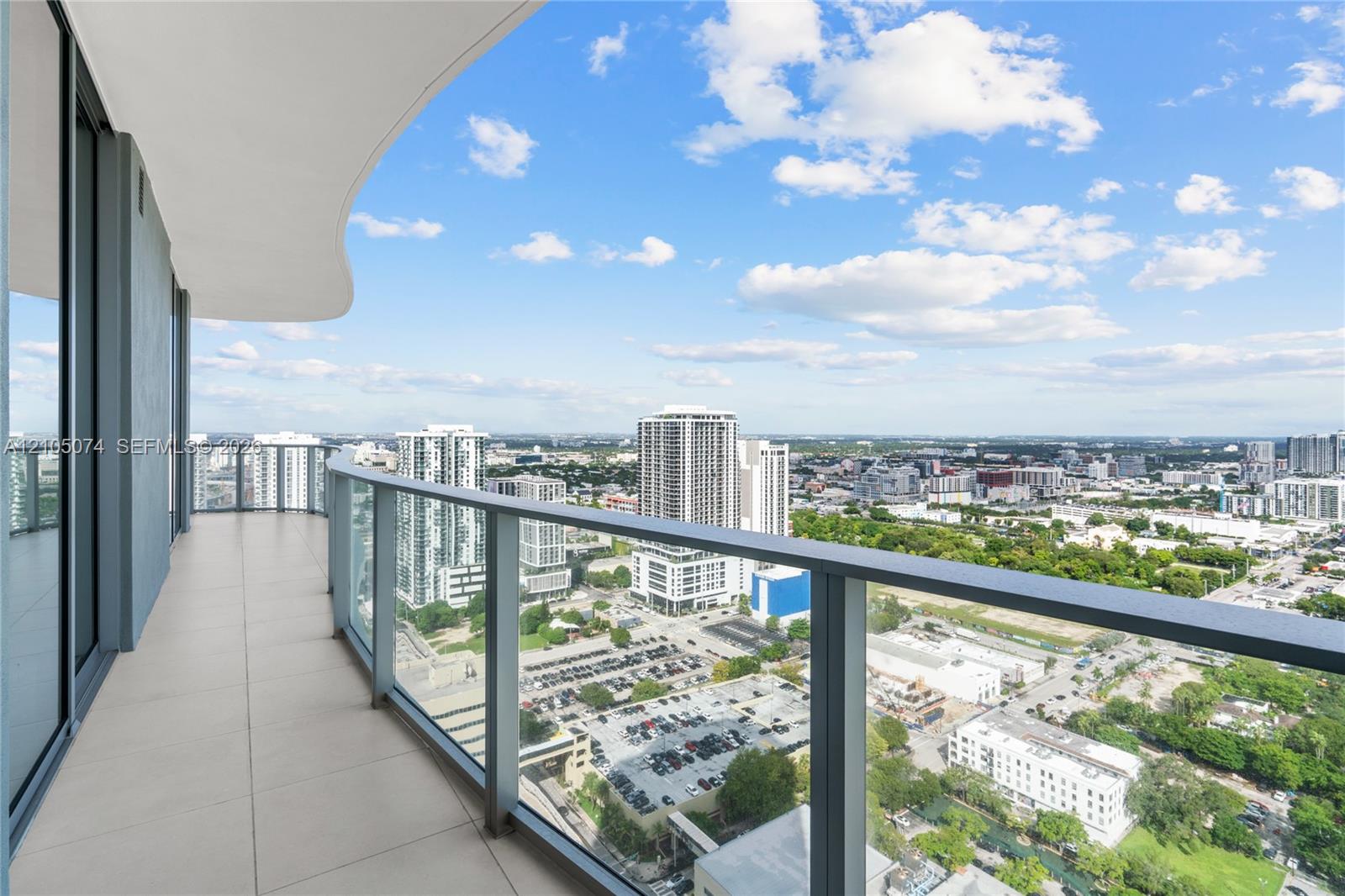 Spacious private balcony featuring large-format tile flooring, a curvilinear overhead architectural element, and a full-height glass railing