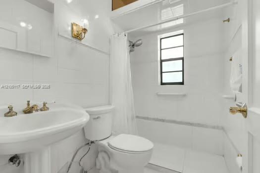 Bathroom featuring a pedestal sink with brass-toned hardware, white wall tile, a combination tub and shower enclosure with a window, and brass-toned sconce lighting