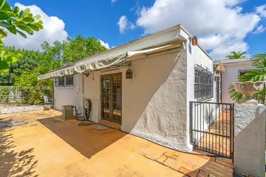 Stucco exterior featuring a retractable awning, glass-paneled double doors, metal security window bars, and a paved patio area