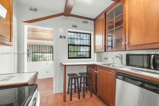 Kitchen featuring wood cabinetry with glass-panel upper doors, integrated breakfast bar, stainless steel dishwasher, built-in microwave, and terracotta tile flooring