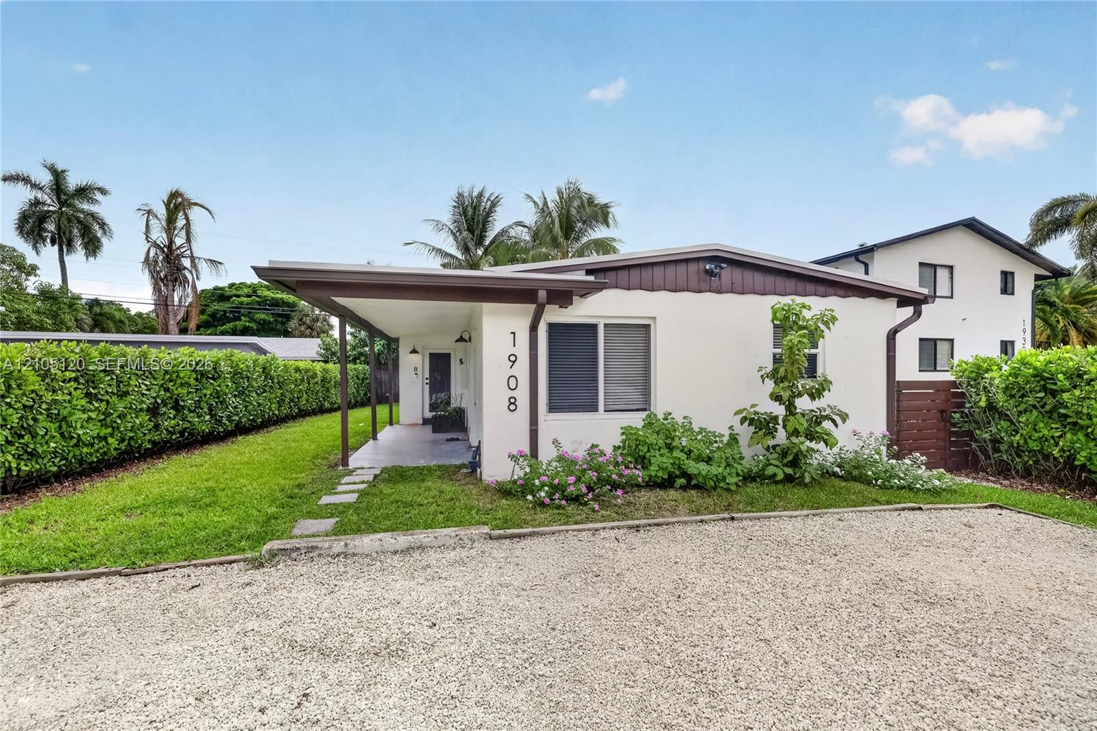 Single-story exterior featuring a covered front porch, light-colored stucco finish, stone-surfaced driveway, and mature perimeter hedging