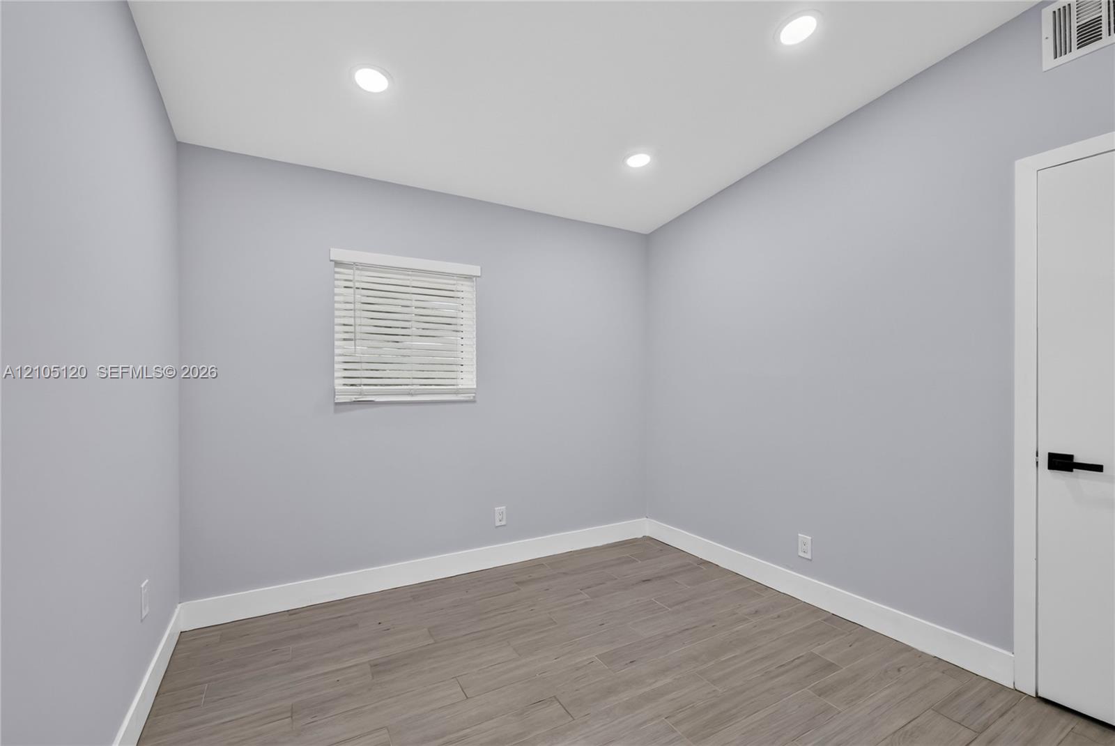 Neutral interior space featuring wood-finish tile flooring, recessed lighting, a single window with horizontal blinds, and a door with matte black hardware