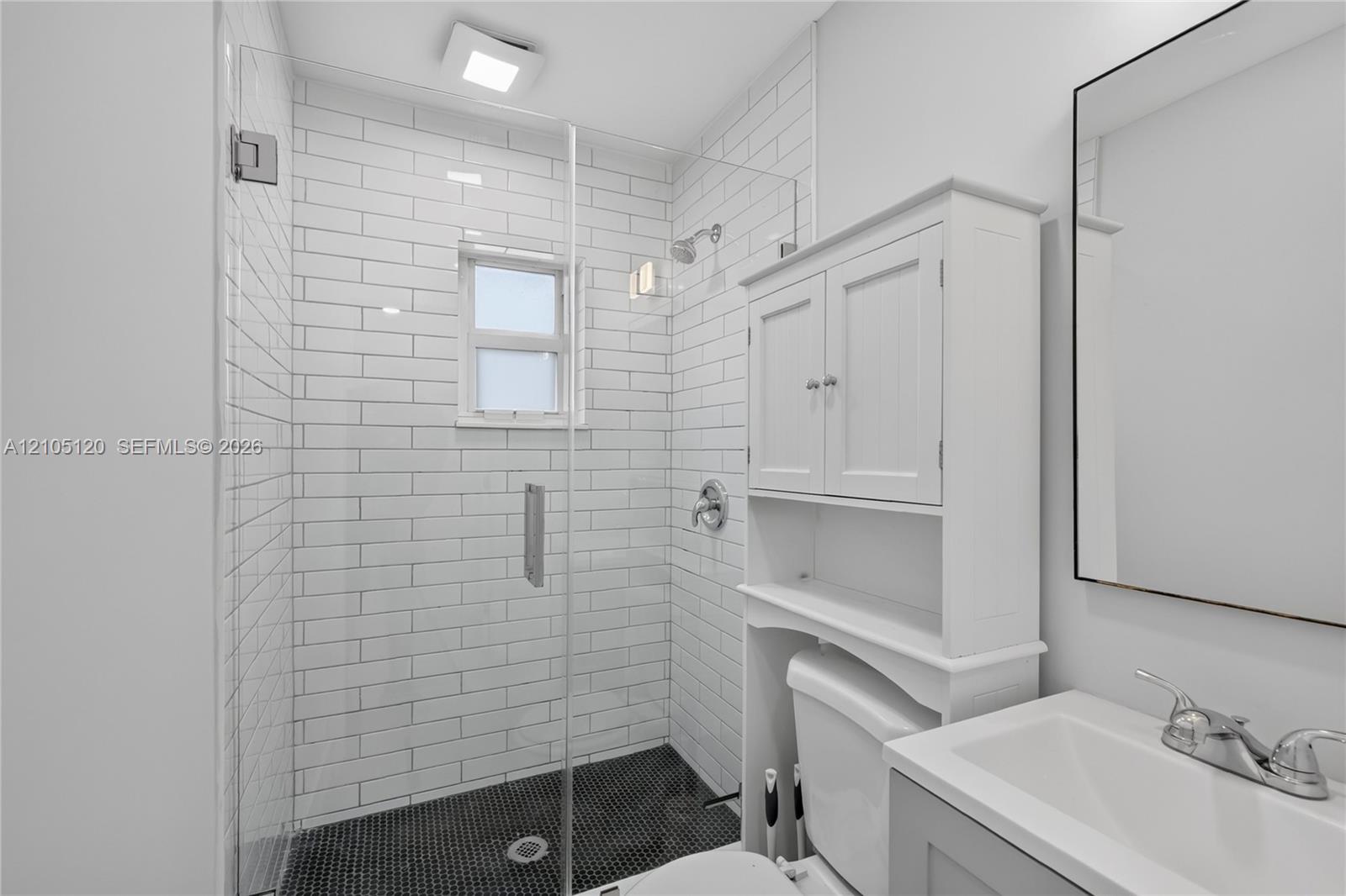 Walk-in shower with white subway tile surround, dark penny tile flooring, and a frameless glass door, accompanied by a white vanity with an integrated sink and an over-the-toilet storage cabinet