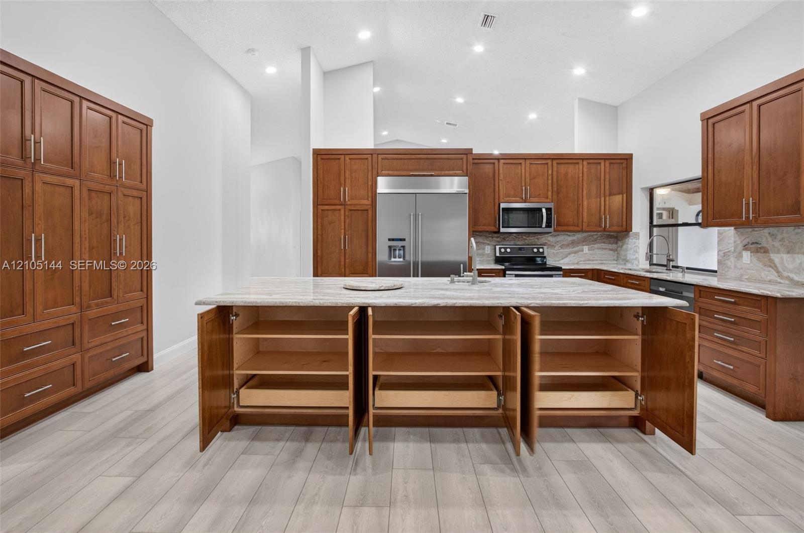Kitchen featuring wood cabinetry, stone countertops, stainless steel appliances, a center island with pull-out shelving, and wood-finish flooring