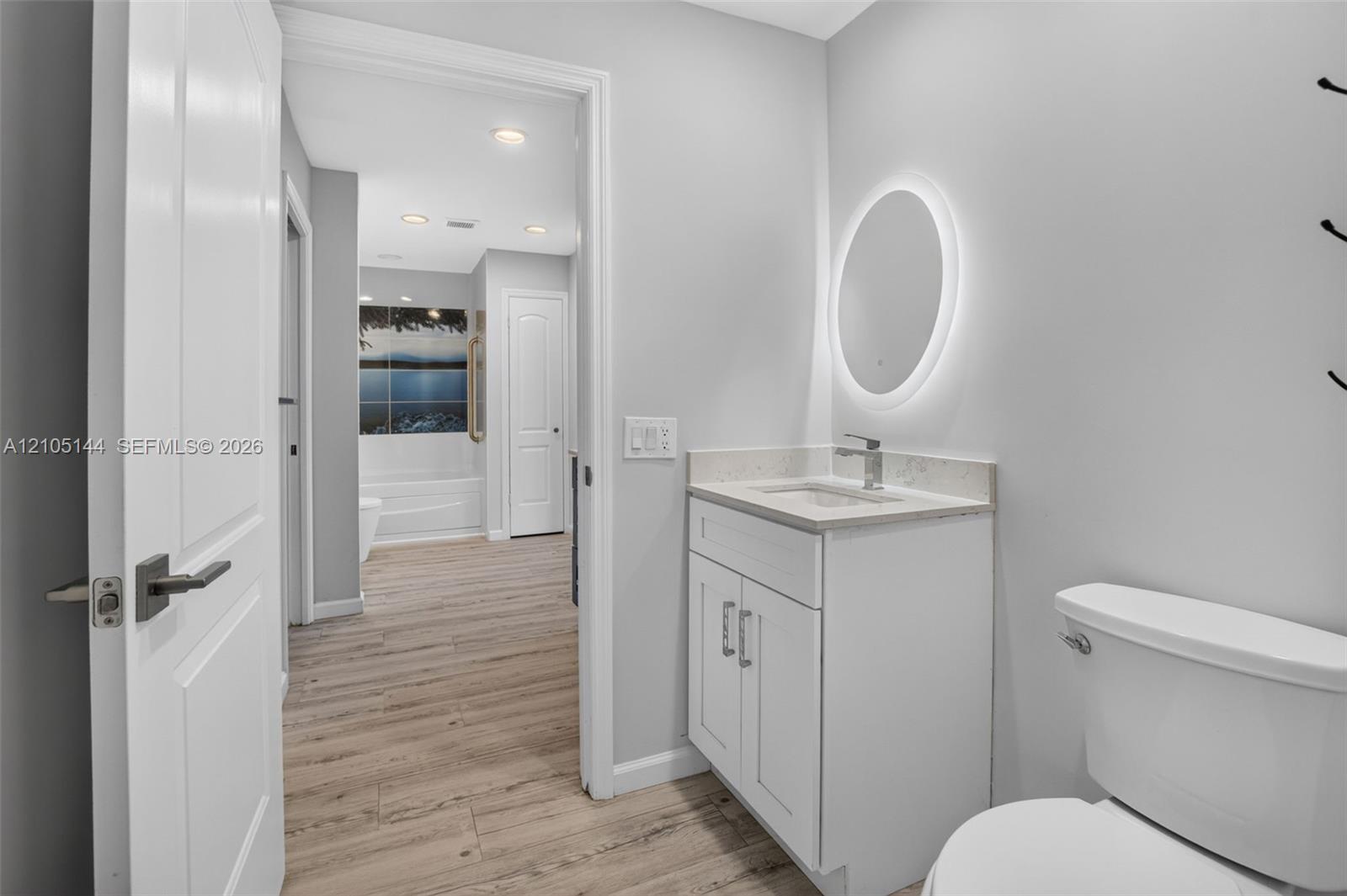 Bathroom featuring a vanity with shaker-style cabinetry, light-toned stone-finish countertop, oval backlit mirror, and wood-finish flooring