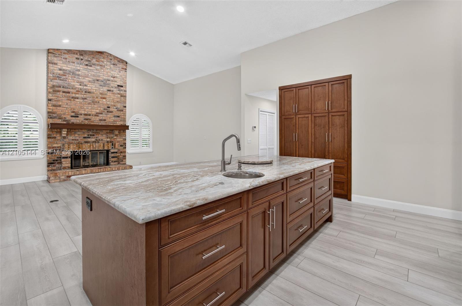Kitchen island with stone-finish countertop, integrated sink, and wood-finish cabinetry, featuring a full-height pantry, wood-finish flooring, and a brick-faced fireplace with a mantel
