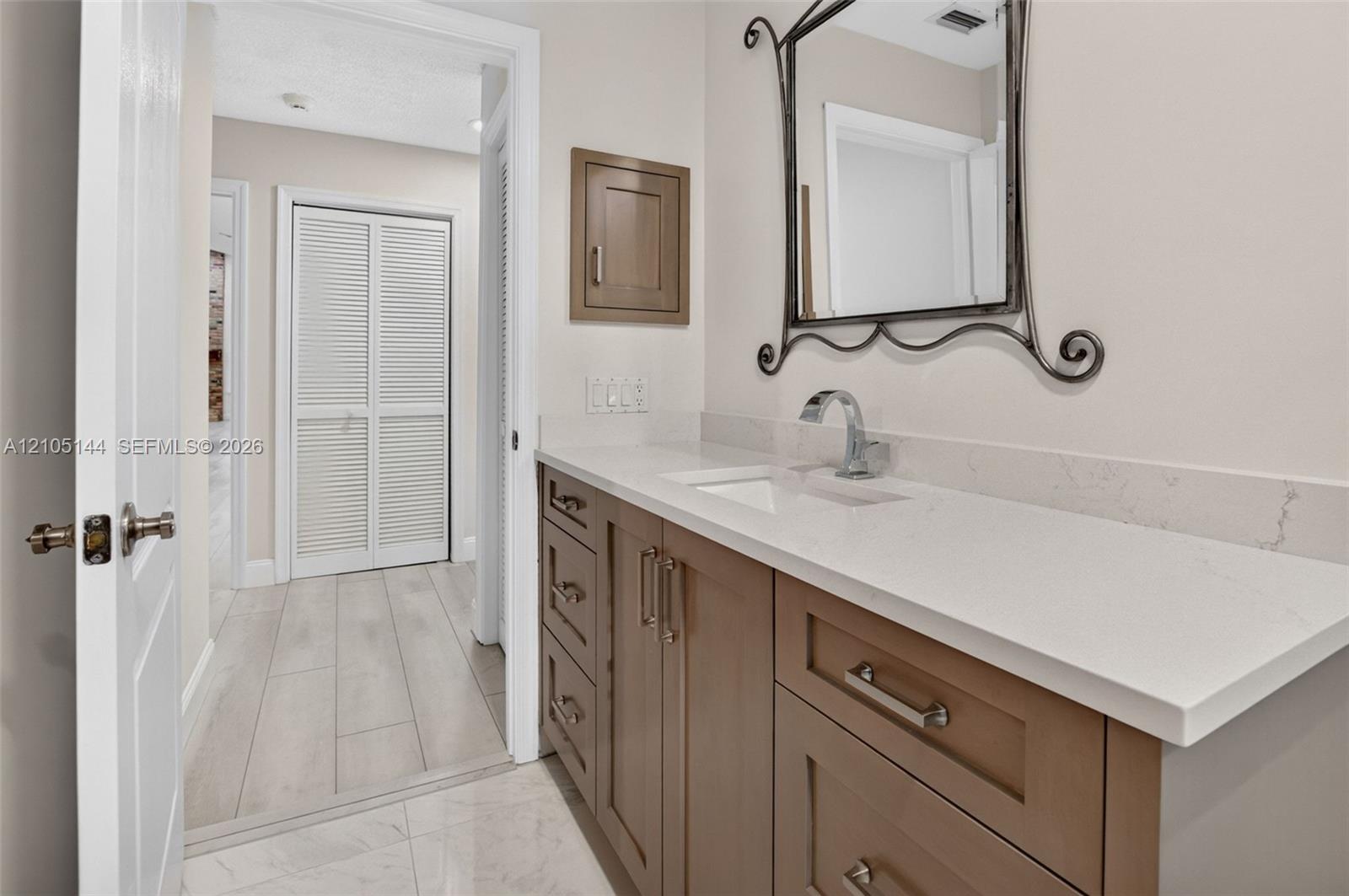 Vanity featuring a stone-composite countertop, undermount sink, shaker-style cabinetry with polished hardware, wall-mounted metallic mirror, and built-in recessed wall cabinet
