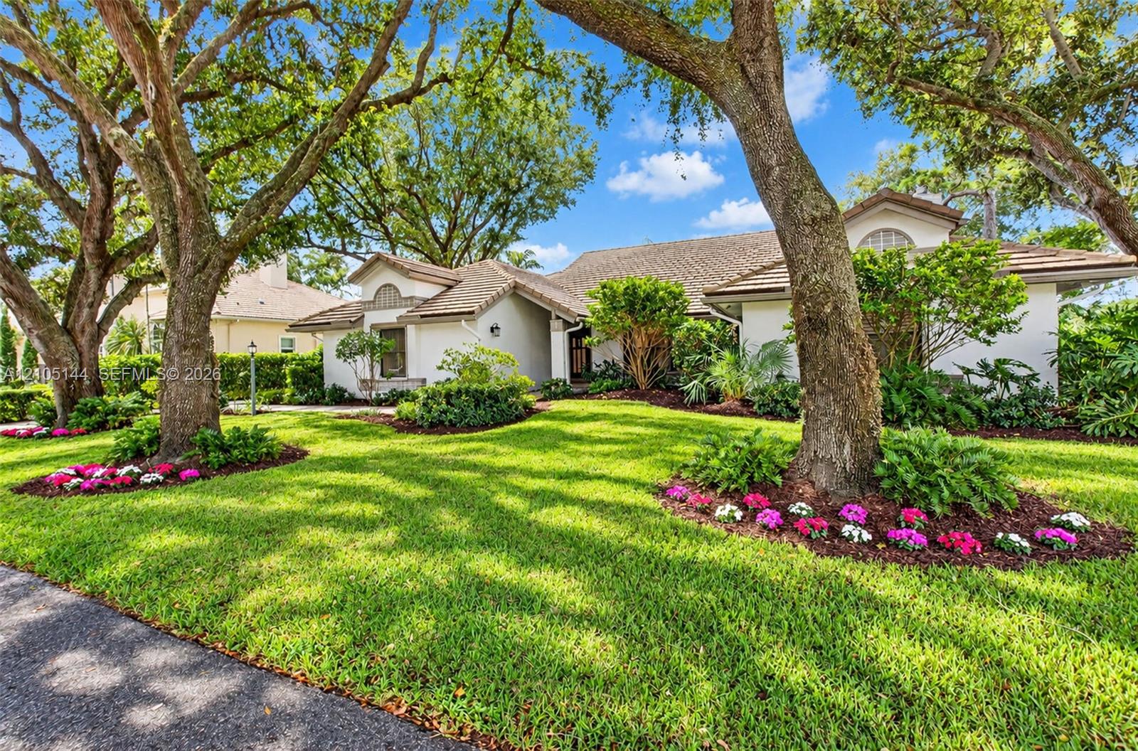 Single-story residence featuring a light-toned stucco exterior, multi-gabled roofline, mature canopy trees, and established landscaping with manicured lawn beds