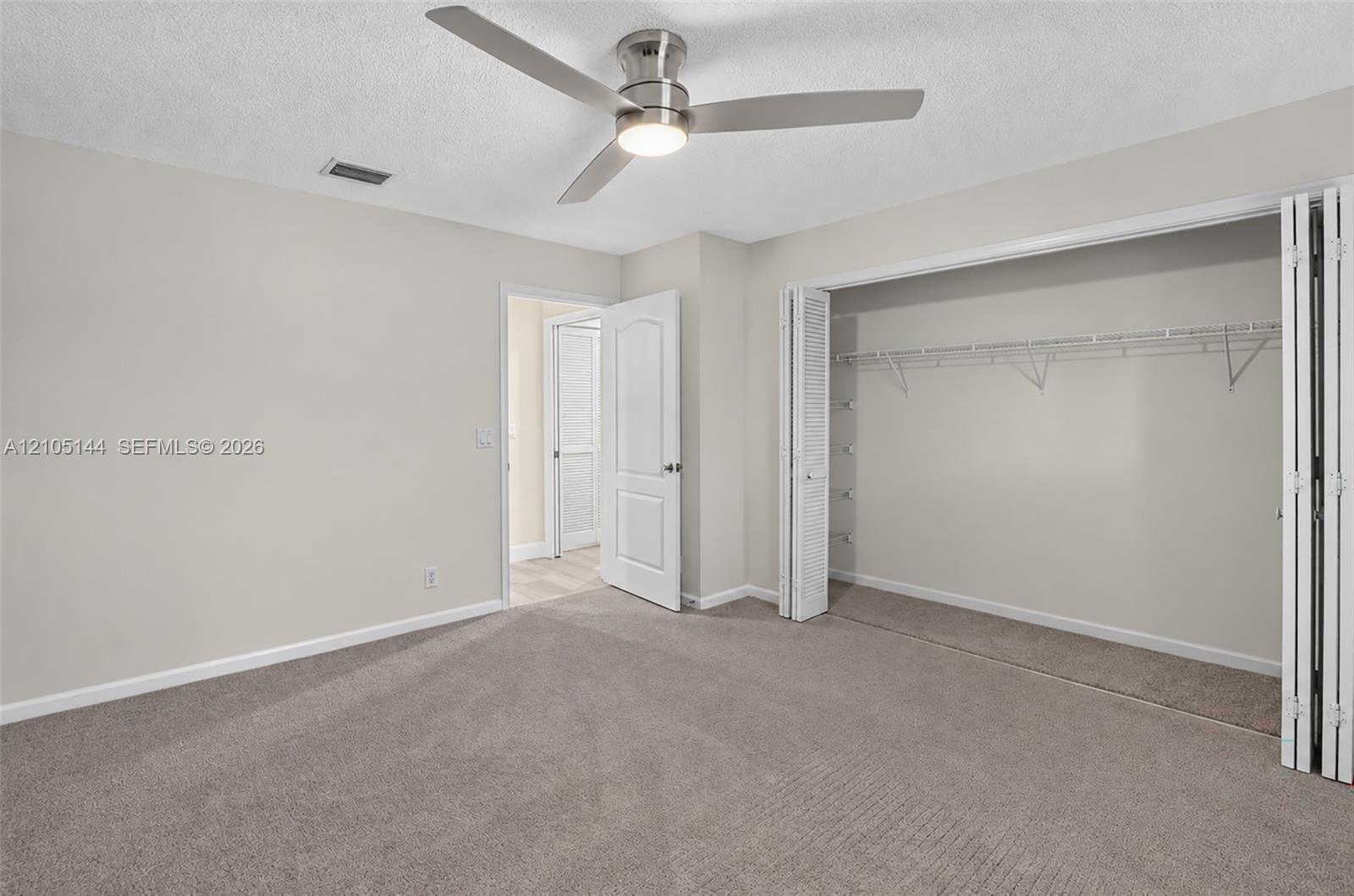 (FAN VIRTUALLY STAGED AS SIMILAR PIECE WILL BE INSTALLED) Neutral-toned interior space featuring neutral wall-to-wall carpeting, a satin-nickel finish ceiling fan with integrated lighting, and a closet with bifold doors and built-in shelving