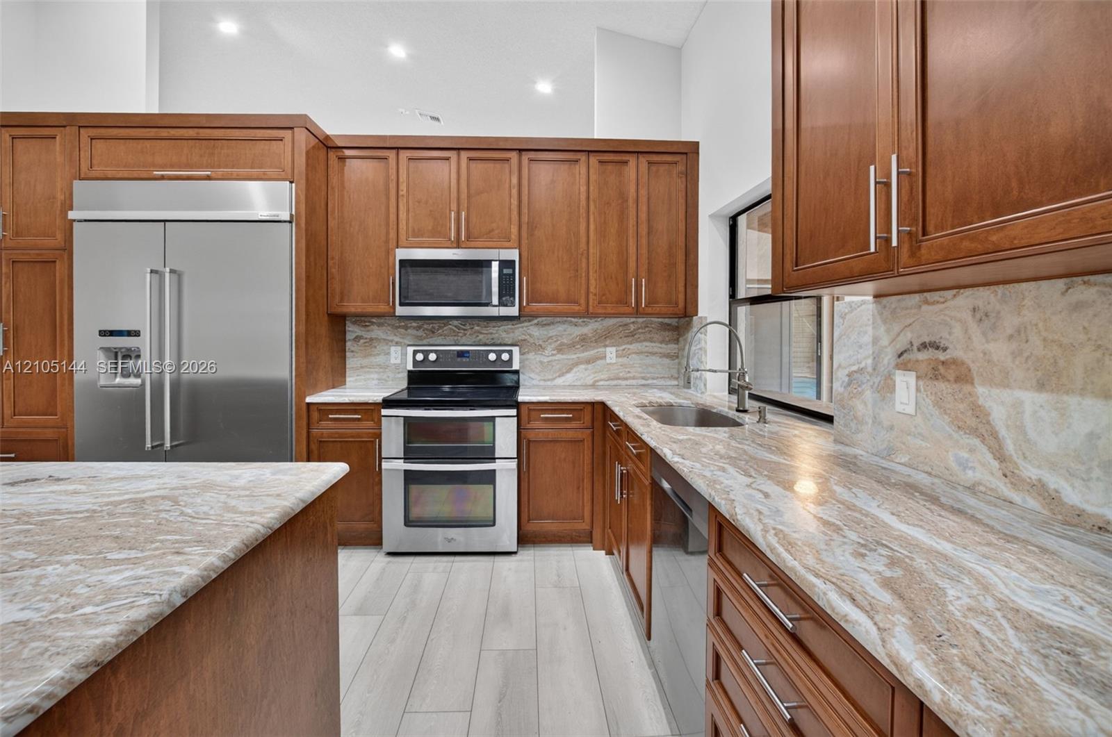 Kitchen featuring wood cabinetry, natural stone countertops and backsplash, stainless steel appliances, and wood-finish flooring