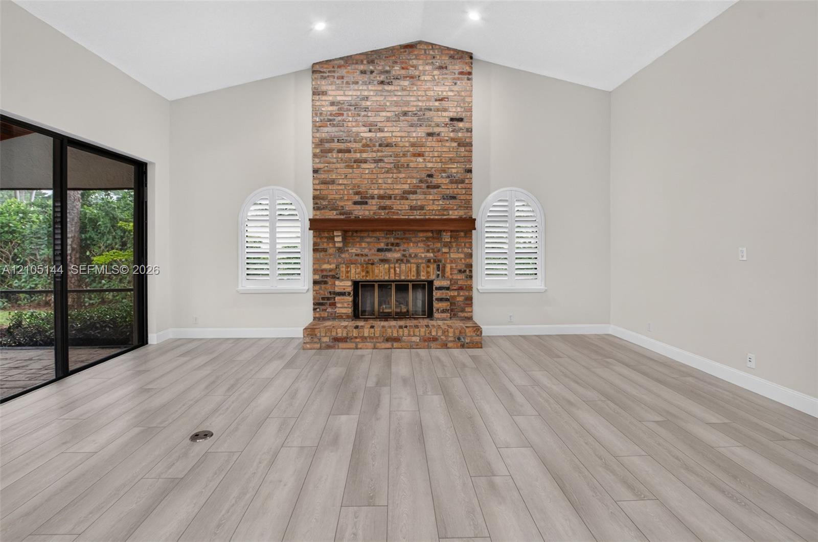 Vaulted ceiling, floor-to-ceiling brick fireplace with a wood mantel, wood-finish flooring, and arched windows with interior shutters